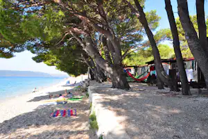 Strandpromenade am Campingplatz mit großen Bäumen, Wohnmobilen und bunten Hängematten, im Hintergrund das Meer.
