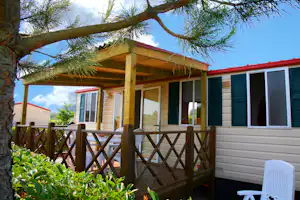 Holzterrasse am Mobilheim mit Markise, Fensterläden und Sitzmöbeln, im Vordergrund ein Baum mit grünen Nadeln.