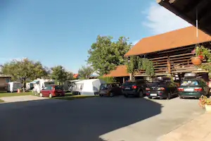Stellplätze mit Wohnmobilen und Zelten, Autos am Rand, Carport mit Ziegeldach und Bäumen im Hintergrund.