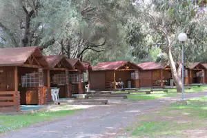 Holzhütten mit Veranda und Sitzbank, davor Tische und Stühle, umgeben von Bäumen auf einer Campinganlage.