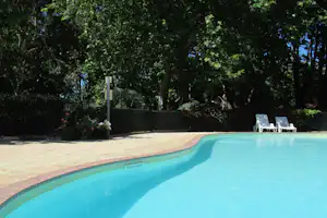 Schwimmbecken mit türkisfarbenem Wasser, gepflasterter Terrasse und Liegestühlen im Schatten von Bäumen und Hecken.