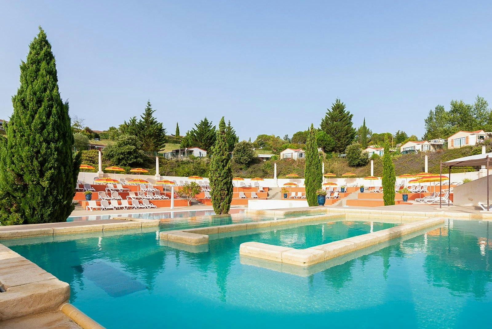 Campingpool mit mehreren Liegen, Sonnenschirmen und gepflegten Hecken, im Hintergrund Reihen von Ferienhäusern.