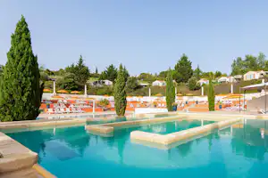 Campingpool mit mehreren Liegen, Sonnenschirmen und gepflegten Hecken, im Hintergrund Reihen von Ferienhäusern.