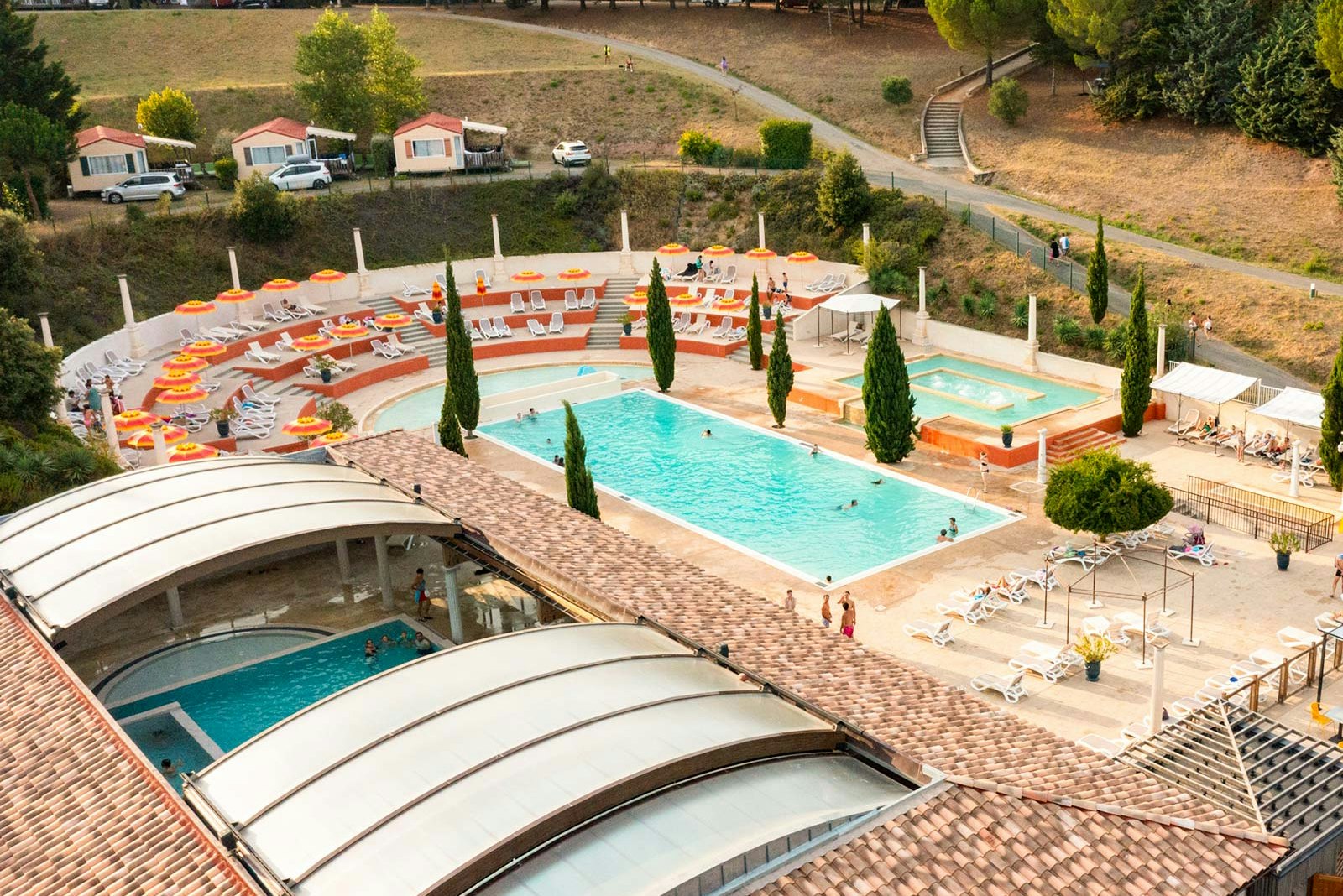 Poolanlage mit zwei Becken, Sonnenliegen und Wohnwagen auf einem Campingplatz, umgeben von Bäumen und Wegen.