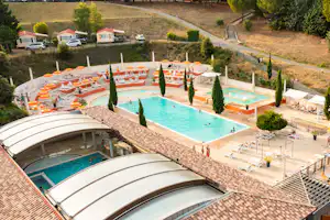 Poolanlage mit zwei Becken, Sonnenliegen und Wohnwagen auf einem Campingplatz, umgeben von Bäumen und Wegen.