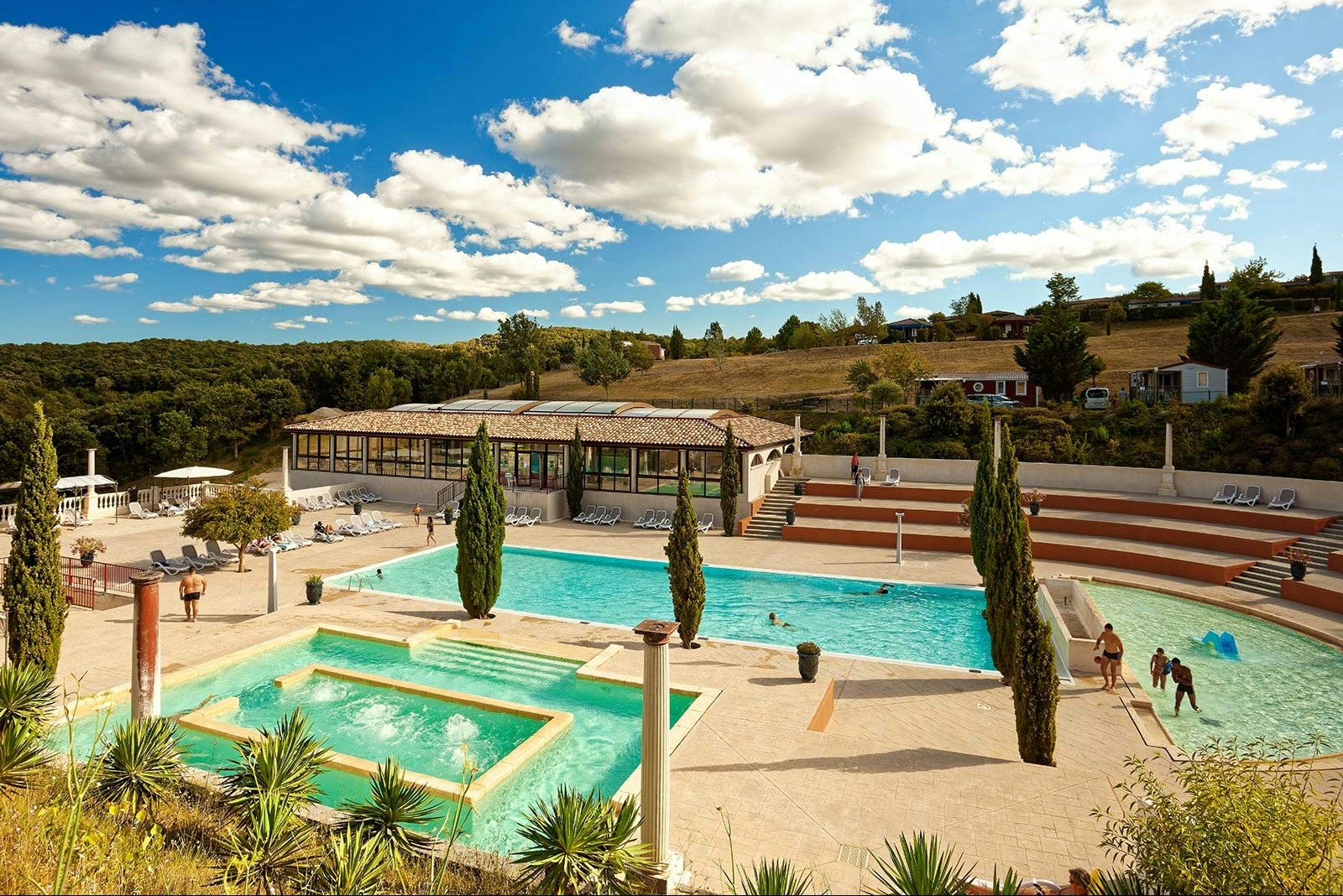 Schwimmbad mit zwei Becken, Liegen und Sonnenschirmen, umgeben von Terrassen, Bäumen und einem Gebäude am Campingplatz.