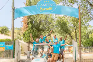Eingangsschild „Camping Paradis“ am Tor, dahinter ein Golfwagen mit mehreren Personen vor Bäumen und Zaun.