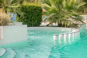 Schwimmbecken mit türkisfarbenem Wasser, Palmen und Hecken hinter einer weißen Mauer am Campingplatz.