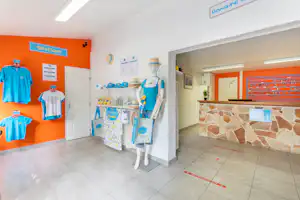 Camping-Shop mit orangefarbener Wand, T-Shirts und Accessoires an der Auslage sowie einer Rezeption im Hintergrund.