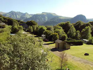 Zelt auf einem Campingplatz auf einer Wiese, umgeben von Bäumen und Wegen, im Hintergrund hohe Berge.