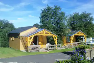 Zwei Holz-Campingunterkünfte mit überdachter Terrasse, Sitzbank und Tisch, umgeben von Rasen und Bäumen.