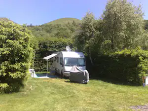 Wohnmobil auf einem Stellplatz im Grünen, mit Vorzelt und Tisch, umgeben von Bäumen und Blick auf grüne Hügel.