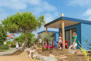 Mobilheim mit Terrasse, davor Spielbereich mit Kindern und Sand, rote Stühle, Bäume und Sträucher am Campingplatzweg.