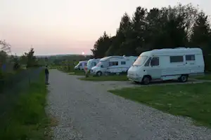 Wohnmobile auf einem Campingplatz entlang eines Kieswegs, mit Rasenfläche und Bäumen am Rand, im Hintergrund eine weite Landschaft.
