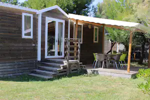 Bungalow mit Holzveranda, Stufen und Terrasse mit Gartenmöbeln vor grünem Rasen und Bäumen.