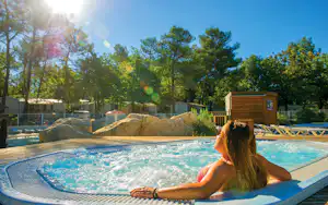 Person am Campingpool im Vordergrund, Holzgebäude und Stellplätze mit Bäumen im Hintergrund.