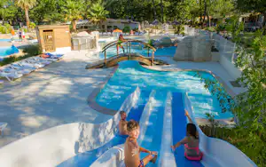 Blauer Pool mit Wasserrutsche und mehreren Becken, umgeben von Bäumen und Campingunterkünften im Hintergrund.