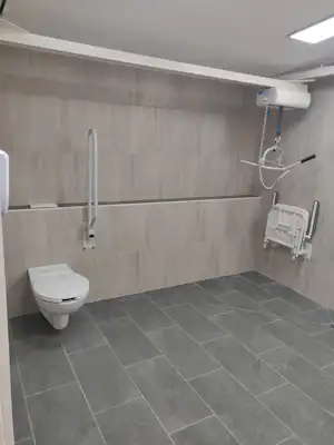 Barrierefreies Badezimmer mit WC, bodenebener Dusche, Haltegriffen und Duschbrause an der Wand, gefliestem Boden.