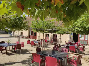 Terrasse mit vielen roten Stühlen und Tischen unter Weinreben, daneben Gebäude und ein Weg auf dem Campingplatz.