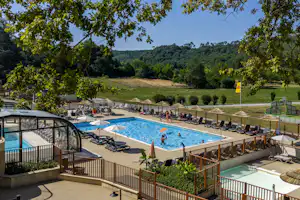 Schwimmbecken mit Liegestühlen und Terrassen am Campingplatz, im Hintergrund Wiesen und bewaldete Hügel.
