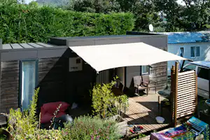 Wohnwagen mit großer Terrasse, Sitzgruppe und Sonnenschirm auf dem Campingplatz, umgeben von Büschen und Bäumen.