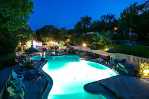 Beleuchteter Campingpool mit Liegen und Sonnenschirmen, umgeben von Bäumen und einem Zaun am Abend.