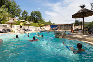 Camping-Pool mit mehreren Personen im Wasser, Sonnenschirmen und Liegen am Beckenrand, im Hintergrund Bäume und Gebäude.