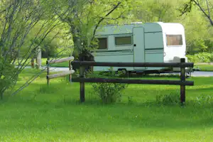 Grüner Wohnwagen auf einem Campingstellplatz, mit Holzbank und Zaun im Vordergrund, umgeben von Bäumen und Rasen.
