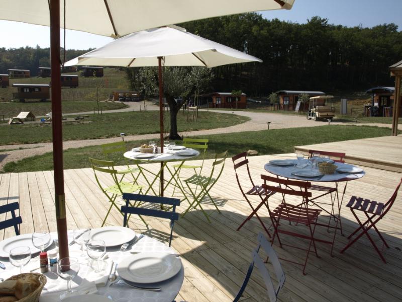 Holzterrasse mit Esstisch unter Sonnenschirm, Campingstühle und Blick auf Wohnwagen und Bungalows im Grünen.