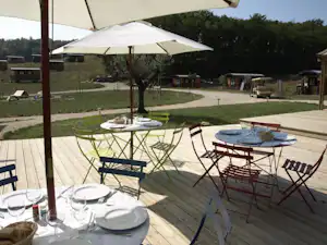 Holzterrasse mit Esstisch unter Sonnenschirm, Campingstühle und Blick auf Wohnwagen und Bungalows im Grünen.