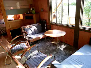 Innenansicht eines Camping-Wohnwagens mit Betten, kleinen Nachttischen und rundem Tisch am Fenster, umgeben von Holzverkleidung.