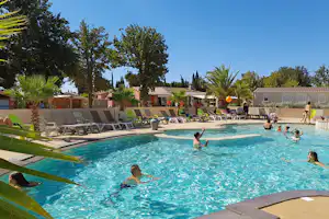 Großer Camping-Pool mit mehreren Liegen, Palmen und Bäumen, daneben ein Gebäude und Personen im Wasser.