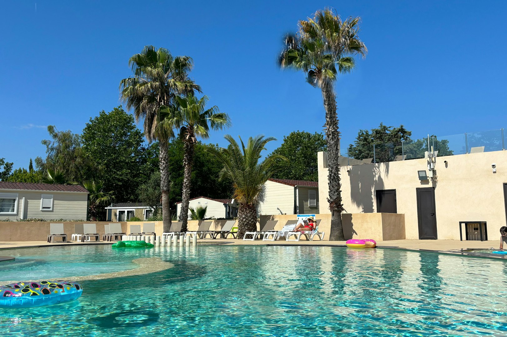 Schwimmbad mit türkisfarbenem Wasser, Liegen und Sonnenschirmen am Rand, Palmen und weißes Gebäude im Hintergrund.