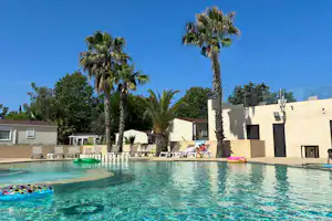 Schwimmbad mit türkisfarbenem Wasser, Liegen und Sonnenschirmen am Rand, Palmen und weißes Gebäude im Hintergrund.