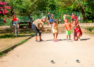 Familie spielt Boule auf einem Campingplatzweg, im Hintergrund Bäume, ein Spielbereich und weitere Personen.