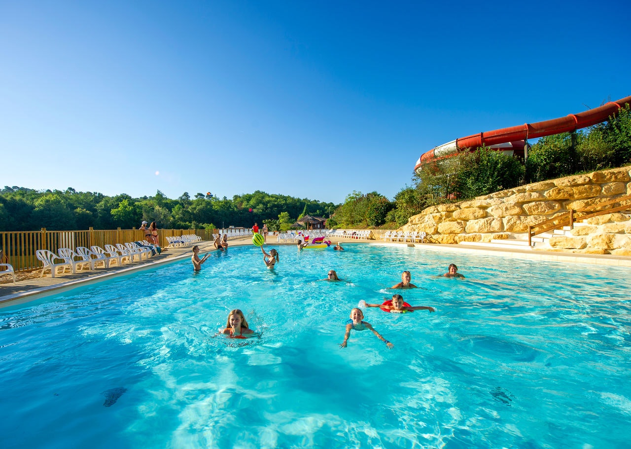 Großes Camping-Schwimmbecken mit vielen Badegästen, Steinmauer und Rutsche, umgeben von Bäumen und blauem Himmel.