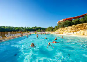 Großes Camping-Schwimmbecken mit vielen Badegästen, Steinmauer und Rutsche, umgeben von Bäumen und blauem Himmel.