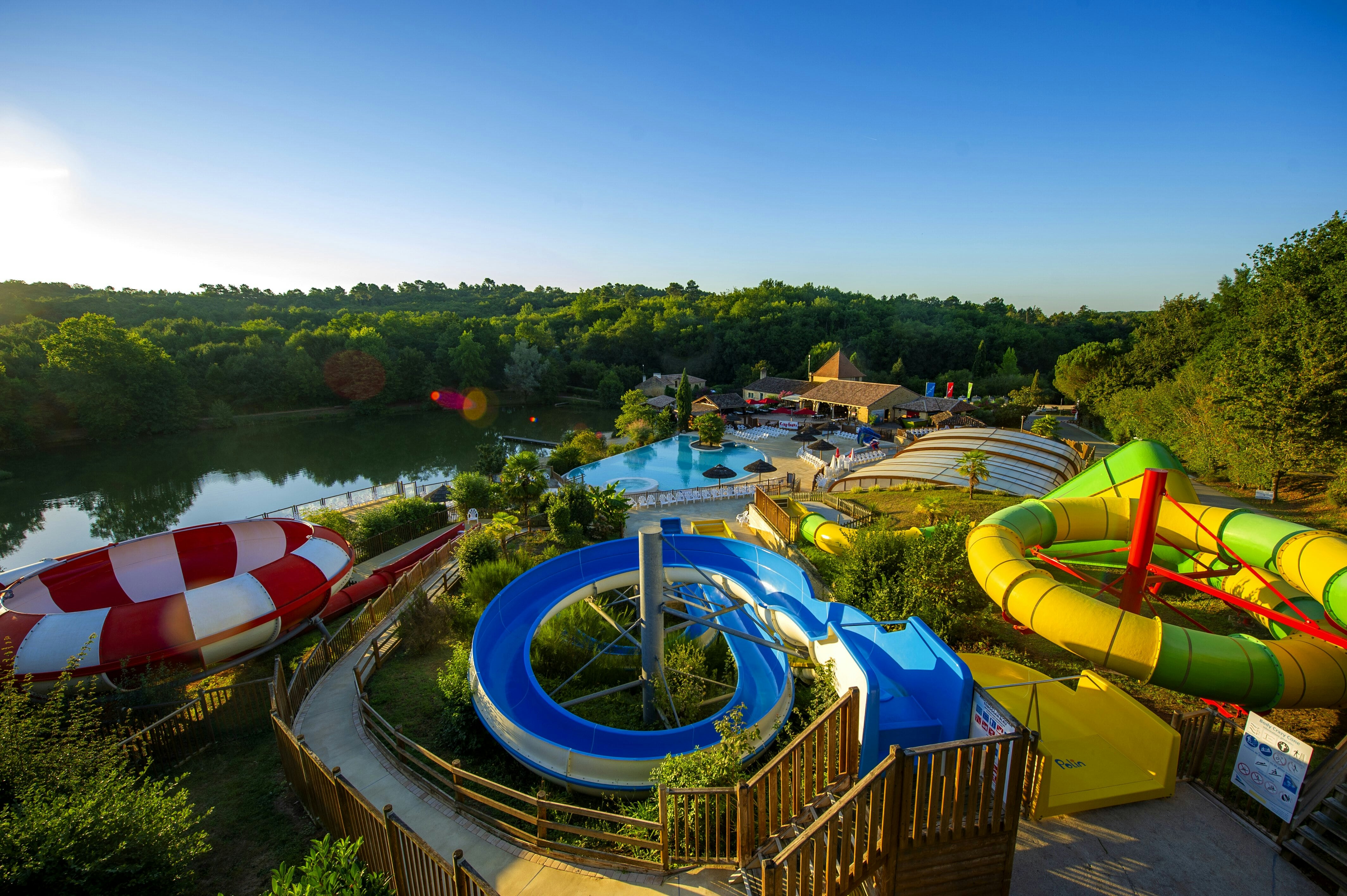 Großer Camping-Pool mit Wasserrutschen, gelben und grünen Rutschen, Liegeflächen und Blick auf den See und Bäume.