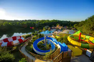 Großer Camping-Pool mit Wasserrutschen, gelben und grünen Rutschen, Liegeflächen und Blick auf den See und Bäume.