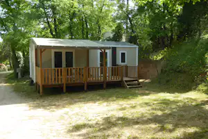 Mobilheim mit Holzveranda und Stufen, umgeben von Bäumen im grünen Campingbereich.