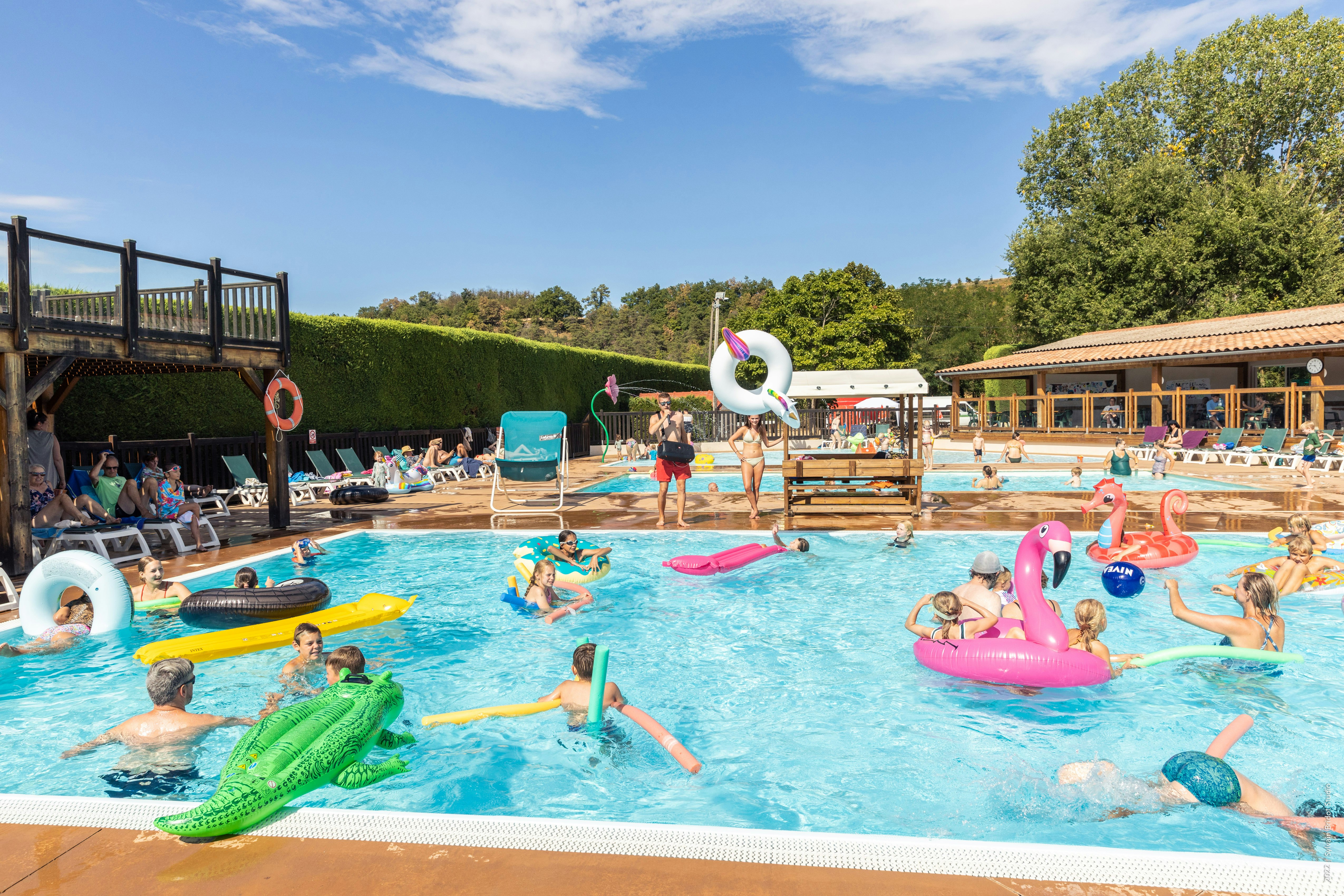 Kinder und Erwachsene im Camping-Pool mit bunten Schwimmtieren, Rutschen und Liegen, im Hintergrund Bäume und Gebäude.
