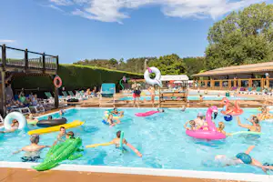 Kinder und Erwachsene im Camping-Pool mit bunten Schwimmtieren, Rutschen und Liegen, im Hintergrund Bäume und Gebäude.