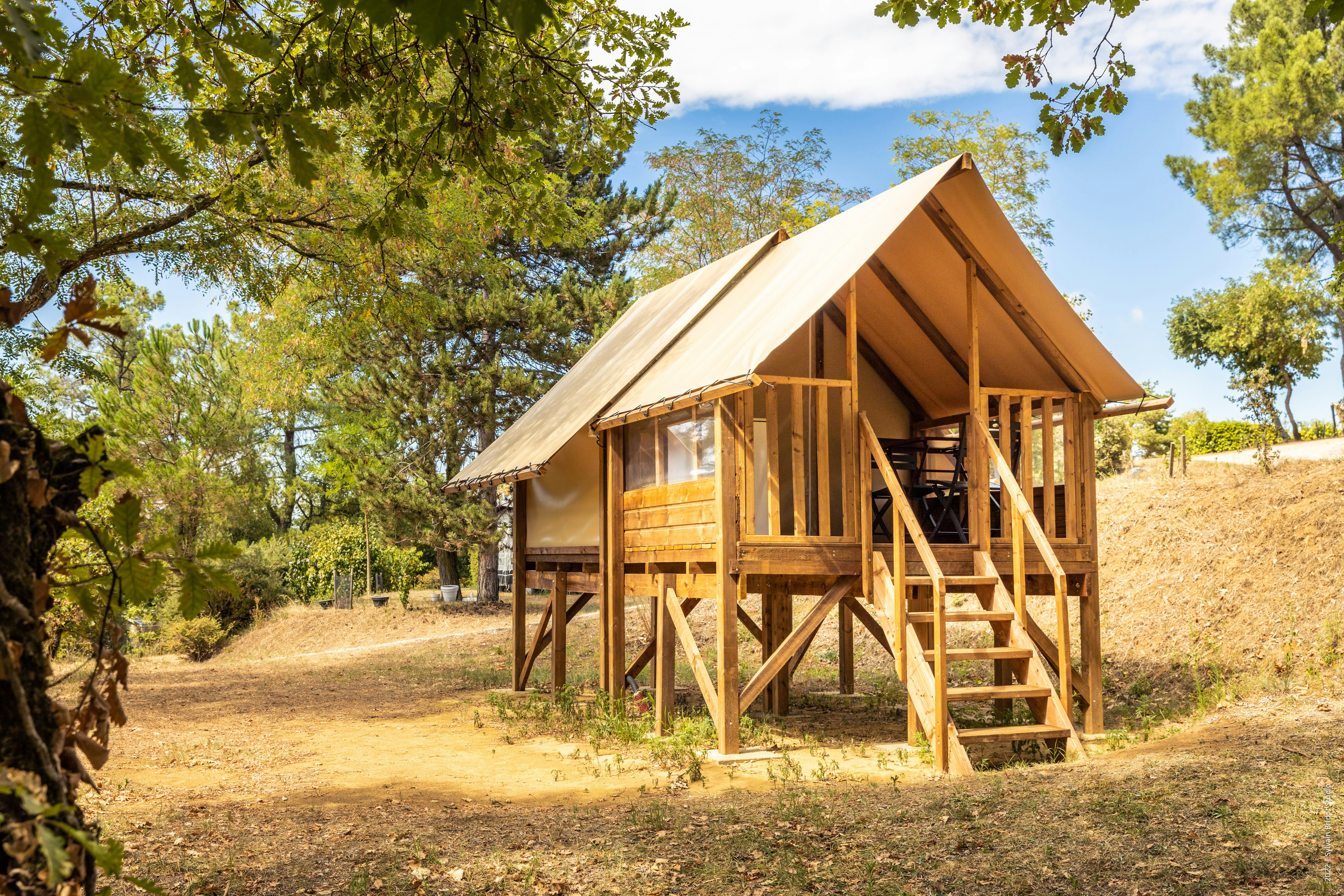 Holzhütte mit Veranda und Außentreppe, umgeben von Bäumen, auf einem grasigen Campingplatz.