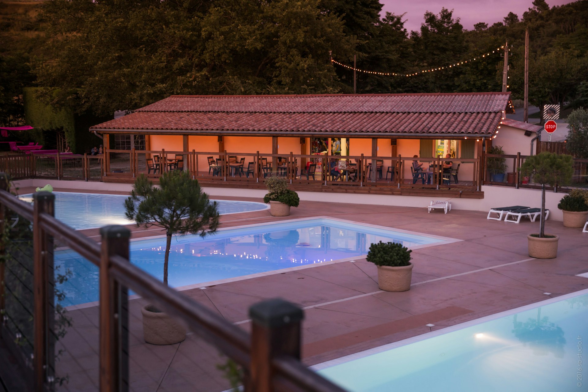 Schwimmbecken mit beleuchteter Terrasse und Liegen am Campingplatz, im Hintergrund ein Gebäude mit rotem Dach und Bäumen.