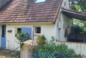 Ferienhaus mit blauem Eingangstürpaar, Ziegeldach und Fenster, davor Steinmauer und Garten mit Sträuchern und hohem Gras.