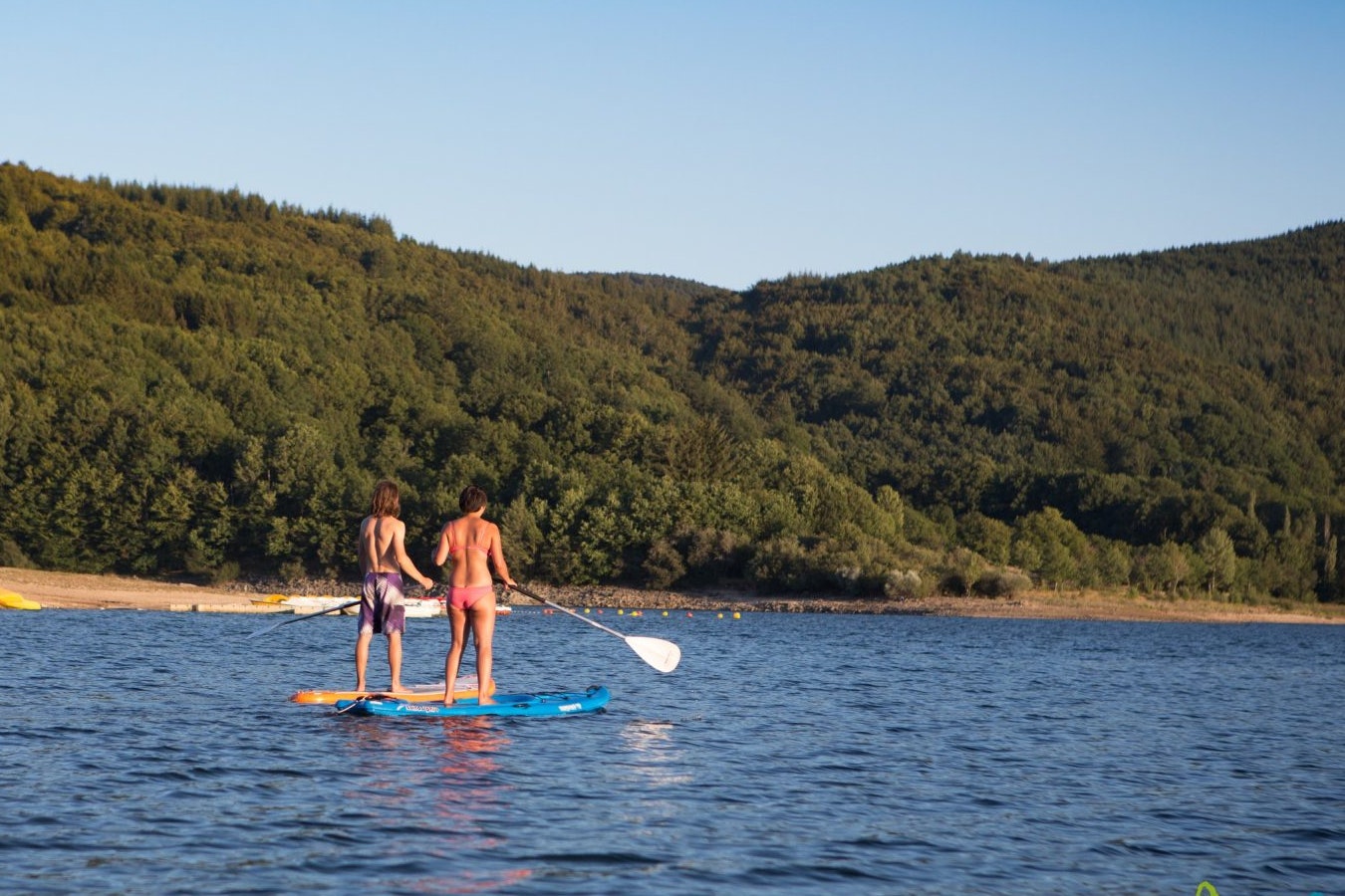 Zwei Personen auf einem SUP-Board paddeln auf einem See, im Hintergrund bewaldete Hügel und ein Paddelboot.