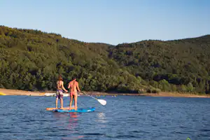 Zwei Personen auf einem SUP-Board paddeln auf einem See, im Hintergrund bewaldete Hügel und ein Paddelboot.