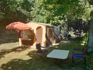 Campingzelt mit Vorzelt und Terrasse, Sonnenschirm, Tisch und Stühlen im Grünen zwischen Bäumen.