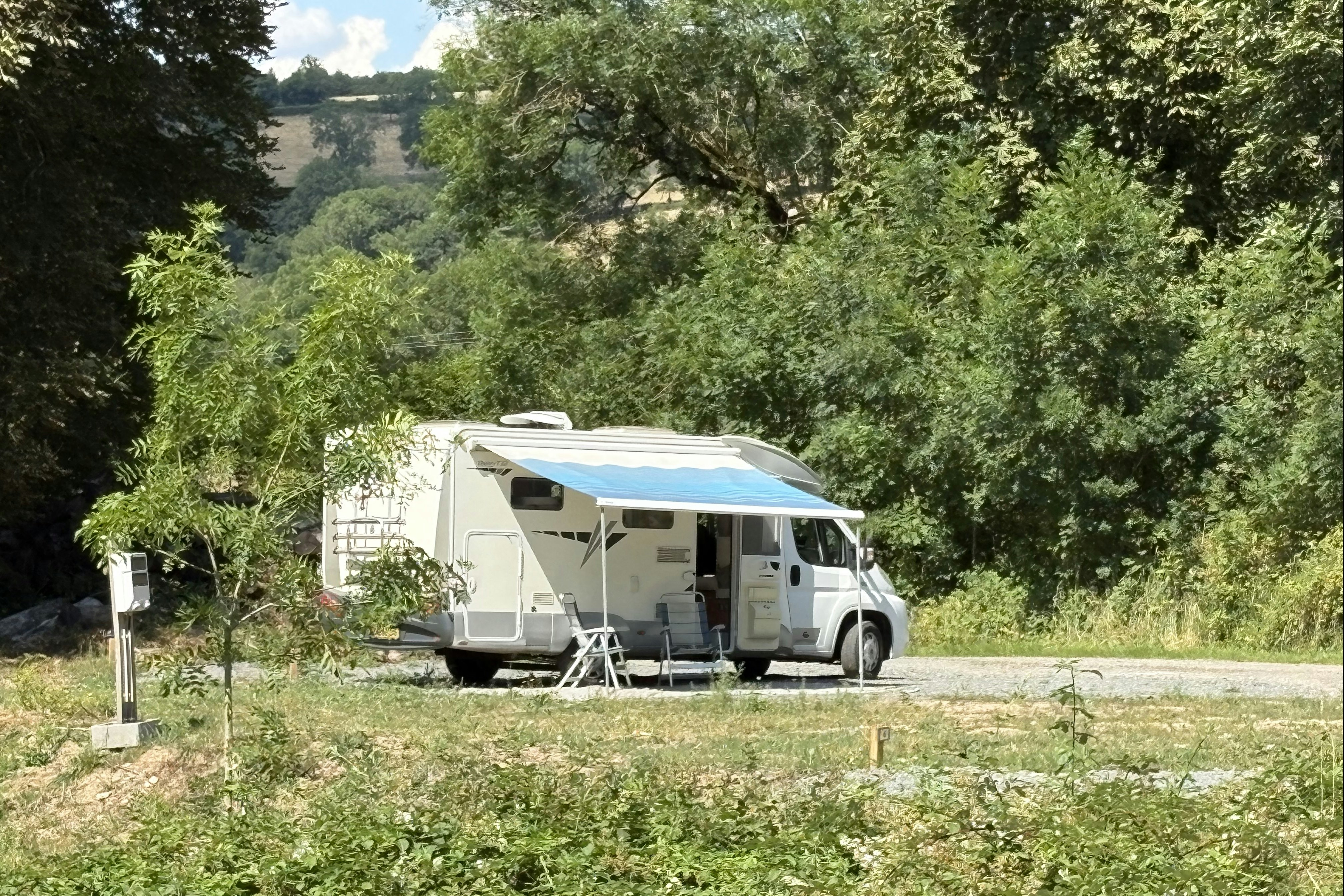 Wohnmobil mit Vorzelt auf einem Campingplatz, umgeben von Bäumen und Grünfläche, mit Tisch und Stühlen am Stellplatz.