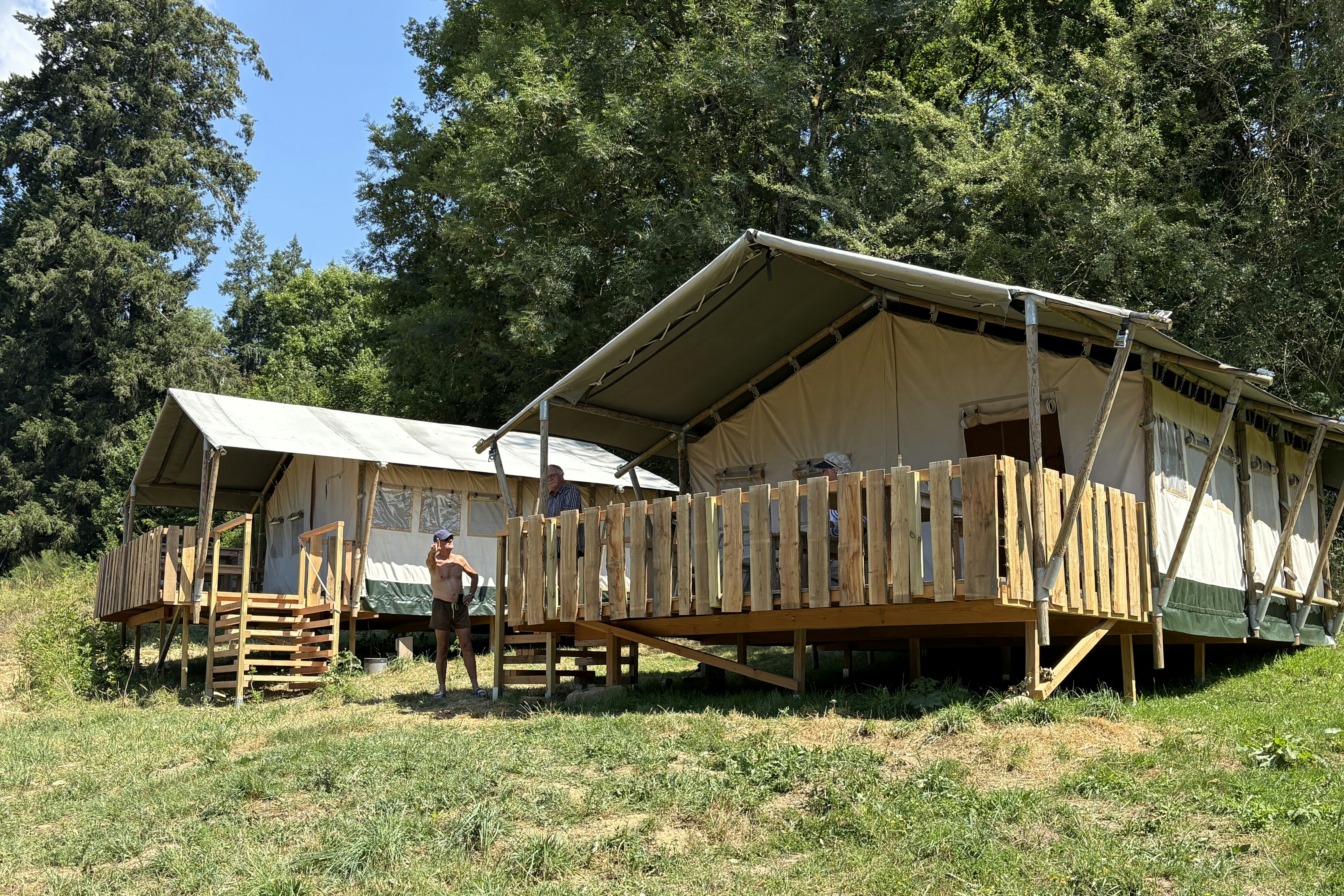 Großes Zelt auf Holzpodest mit Geländer, daneben eine Person vor dem Eingang, umgeben von Bäumen und Wiese.